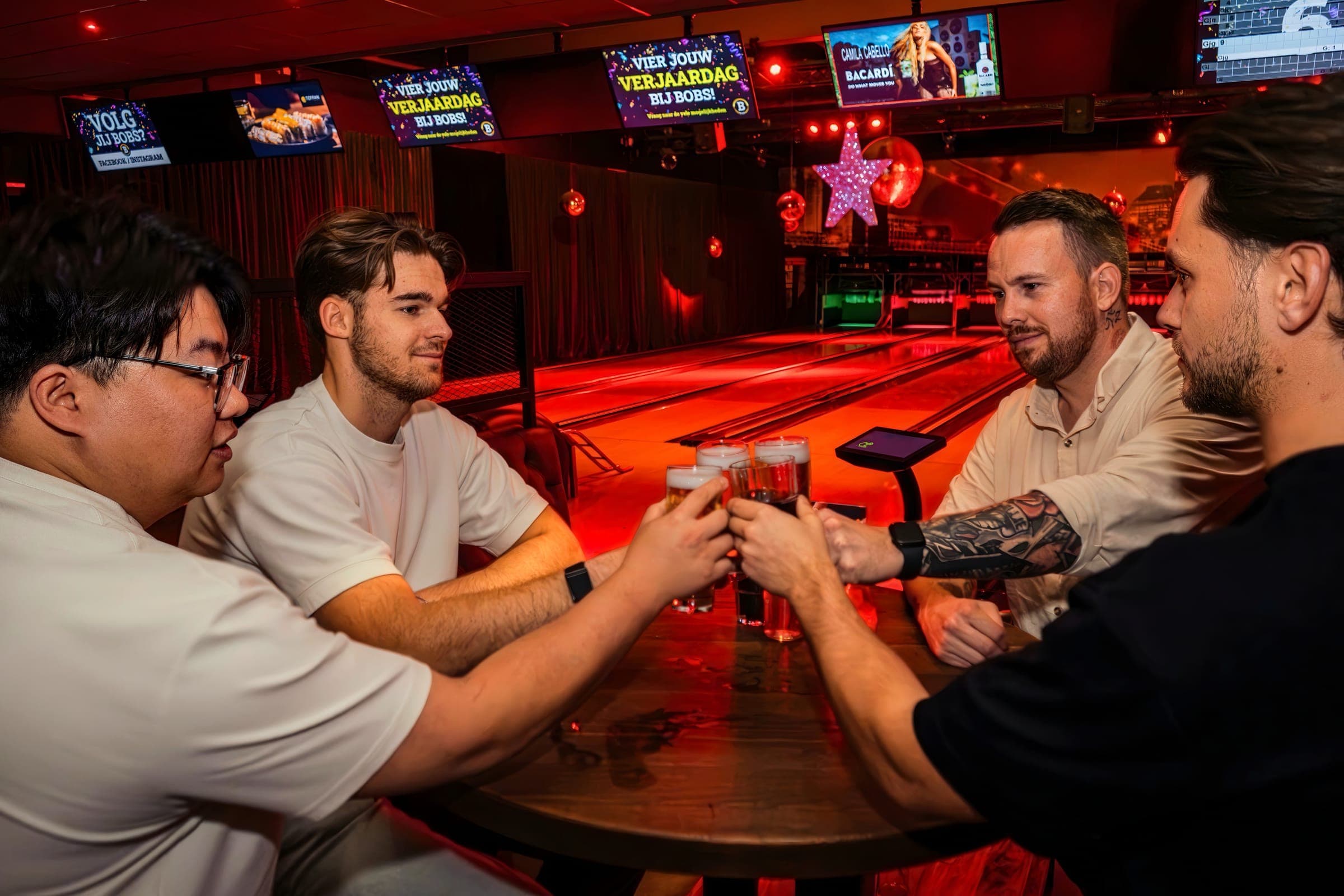 Vier vrienden proosten met bier aan een tafel naast de bowlingbanen, onder rood licht